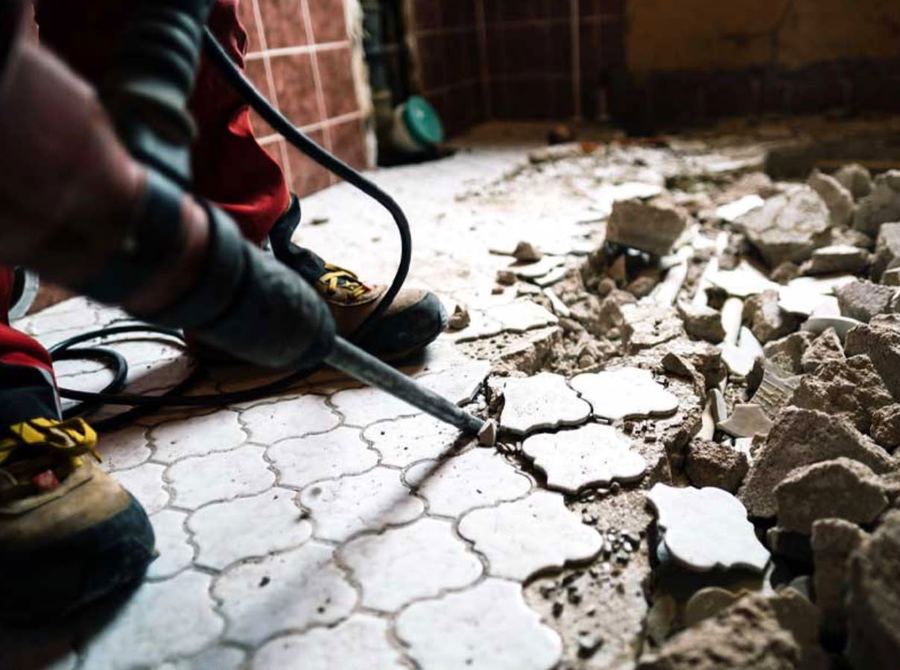 Handwerker stemmt mit dem Abbruchhammer alte weiße Bodenfliesen heraus, daneben liegt bereits Fliesen- und Estrichschutt.