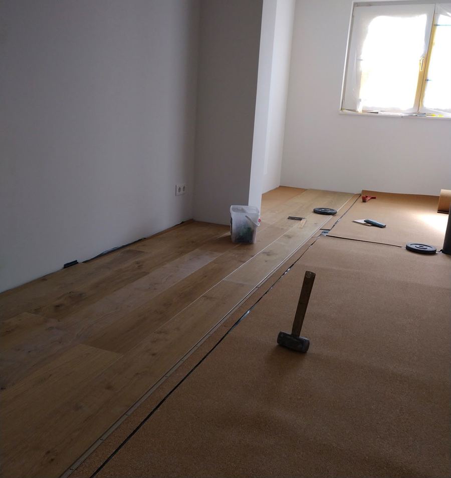 Verlegebeginn von Landhausdielen auf brauner Dämmunterlage, daneben Gummihammer, Eimer und Beschwerungsgewichte.