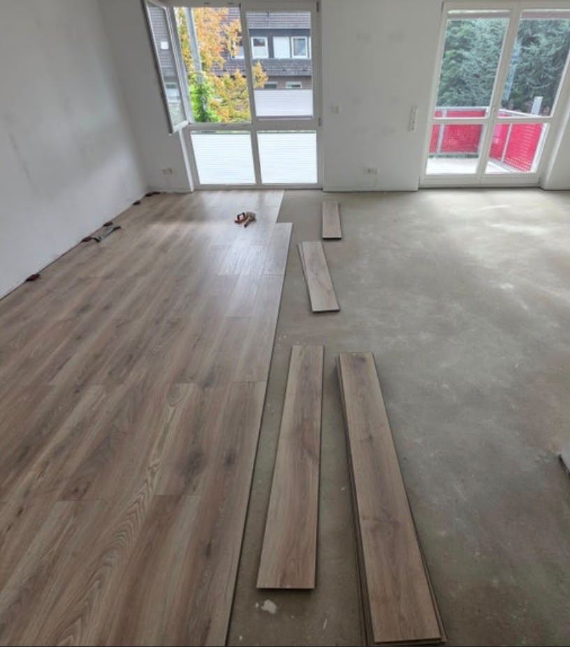 Halb verlegter Laminatboden in Eiche-Optik in einem leeren Wohnzimmer mit bodentiefen Fenstern, einzelne Dielen liegen zum Verlegen bereit.