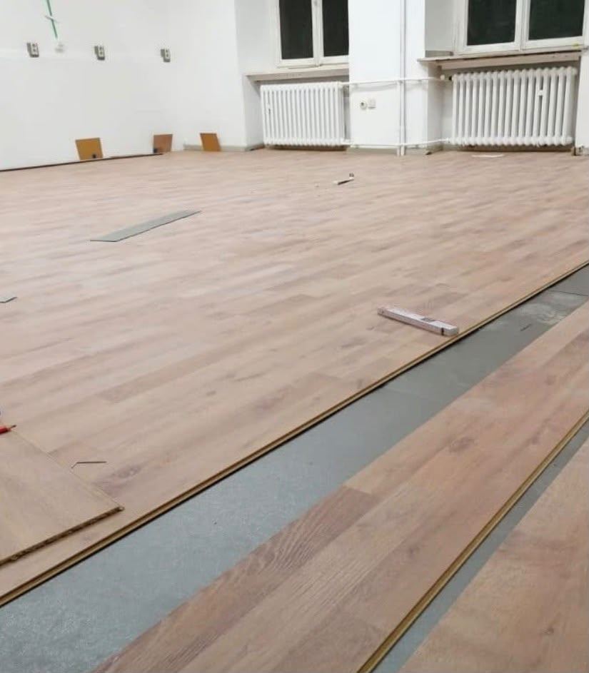 Großflächige Verlegung von hellem Vinyl-Klickboden in einem Altbauraum mit Heizkörpern, im Vordergrund noch offene Reihen auf der Trittschalldämmung.