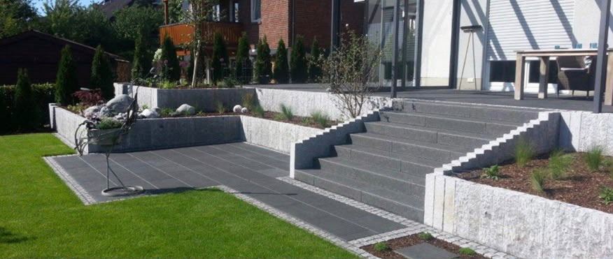 Fertige Gartenanlage mit Granittreppe, dunkler Plattenterrasse, Natursteinmauern und angelegtem Rasen am Haus.