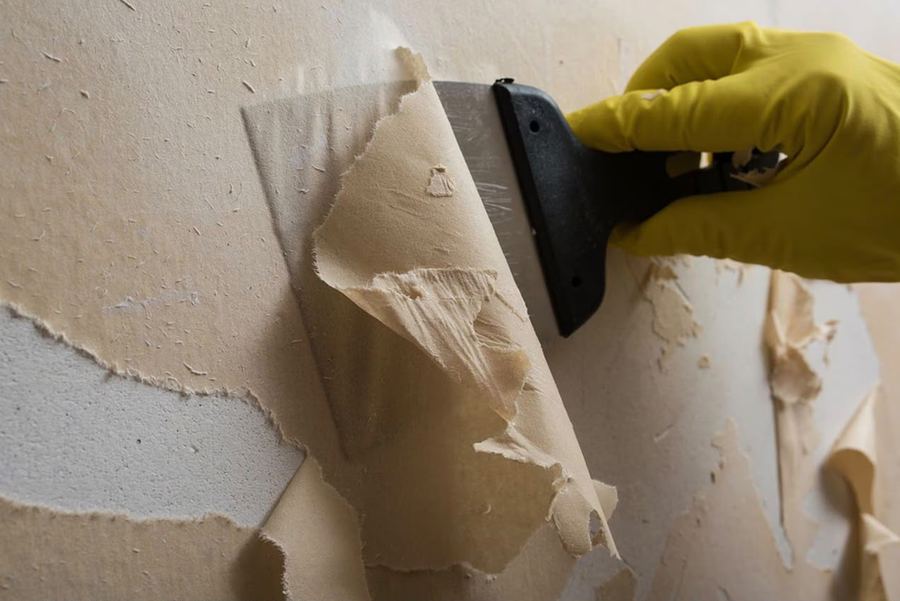 Hand im gelben Arbeitshandschuh zieht mit einem Spachtel alte Tapete von der Wand ab, daneben bereits freigelegter Putz.