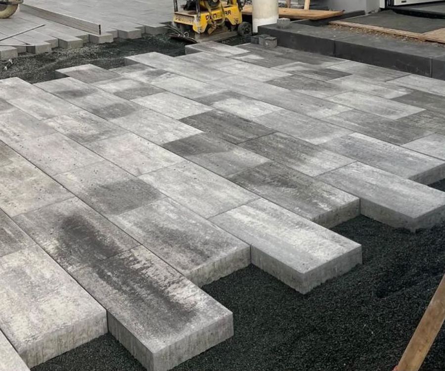 Großformatige Betonplatten werden im Läuferverband auf Splitt verlegt, im Hintergrund steht eine Rüttelplatte.