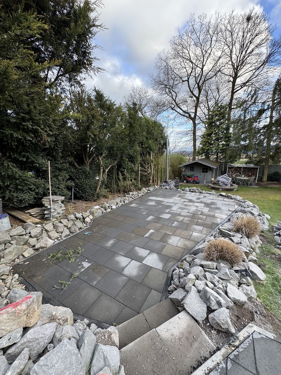 Neu angelegte Gartenterrasse aus grauen Platten, eingefasst mit Natursteinen und Gräsern.