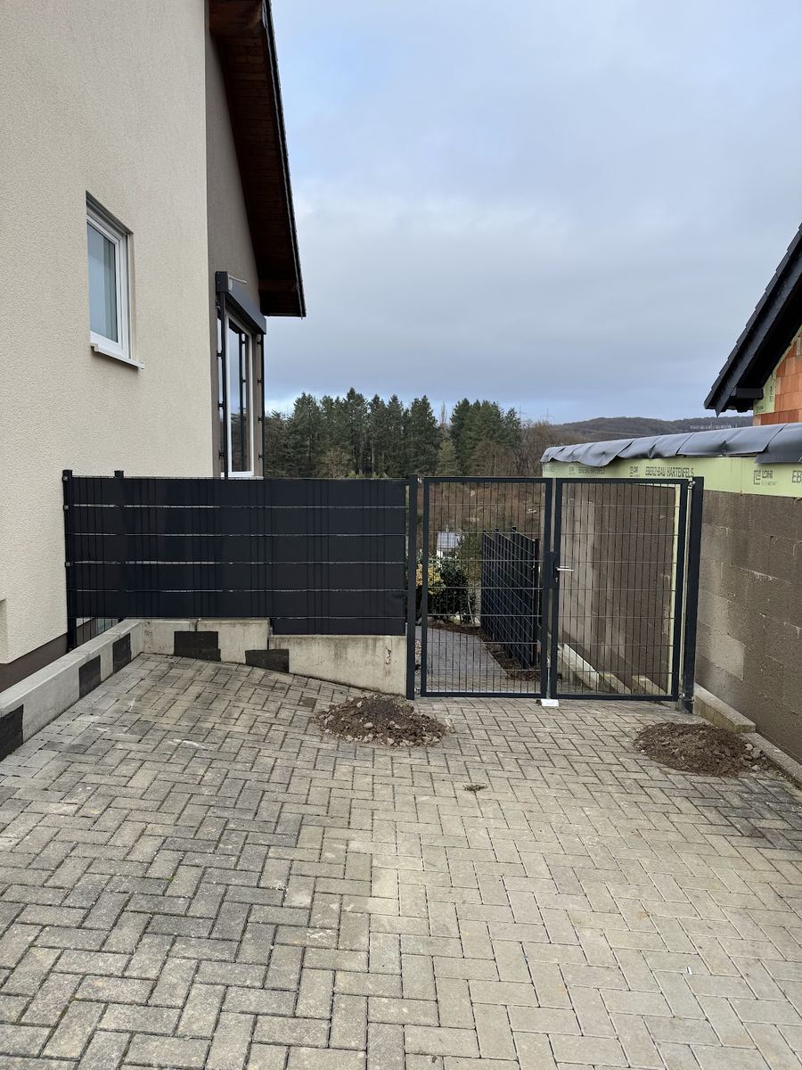 Gepflasterter Hof mit neu montiertem anthrazitfarbenem Gartentor und Sichtschutzzaun am Neubau.