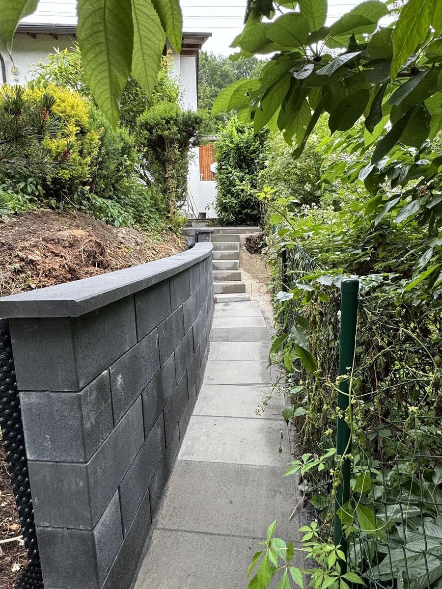 Fertige anthrazitfarbene Blockstein-Stützmauer mit Abdeckplatten entlang eines neuen Plattenwegs mit Treppe im Grünen.